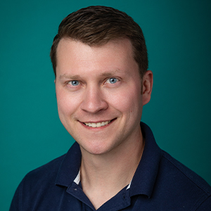 Male physical therapist smiling in front of blue photo backdrop.