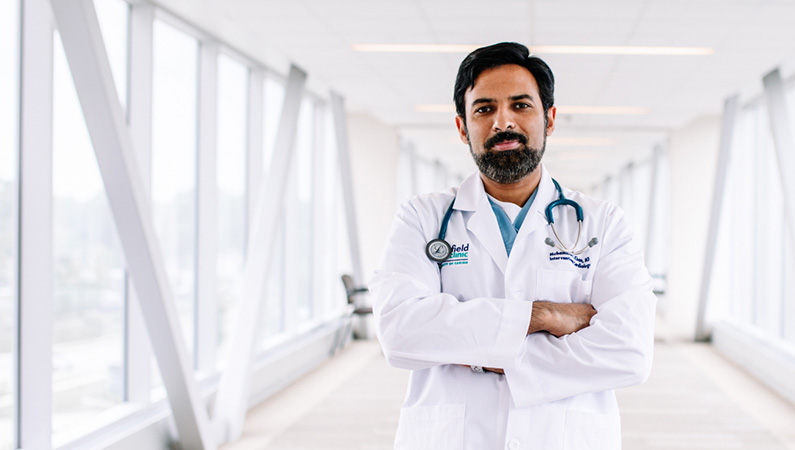 Man wearing a white doctors coat folding arms and smiling in a well-lit walkway