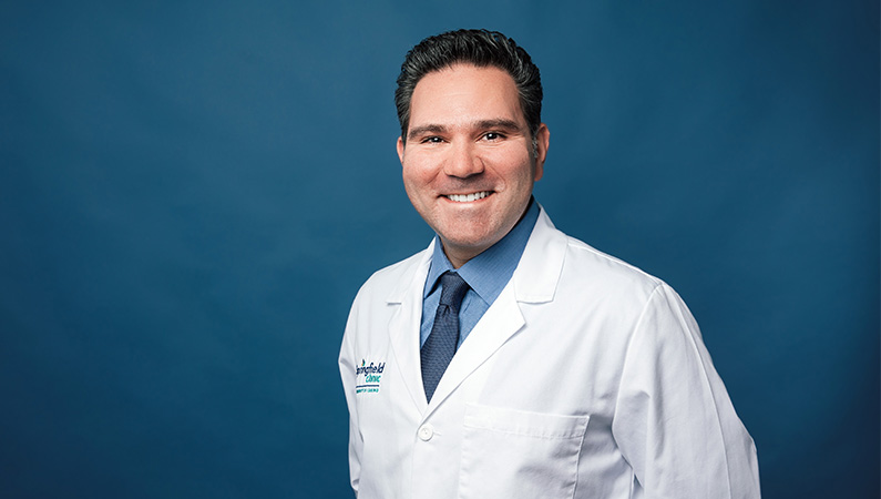 Male doctor in white doctor coat smiling in front of blue photo backdrop.
