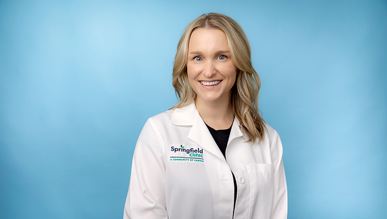Female nurse practitioner in white coat smiling in front of light blue backdrop.