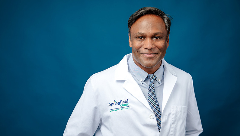 Male neurology doctor in white lab coat smiling in front of navy blue backdrop.