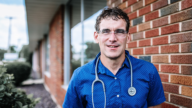 Man wearing a stethoscope around his neck, standing outside of the doctors office building.