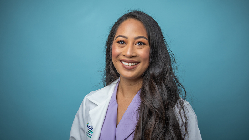 Female nurse practitioner in white medical coat smiling in front of light blue photo backdrop.