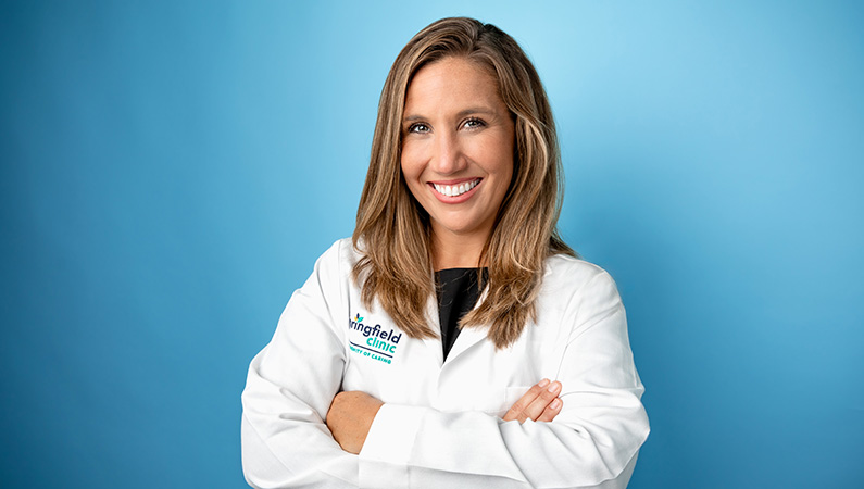 Female physician assistant in white lab coat smiling in front of a light blue photo backdrop.