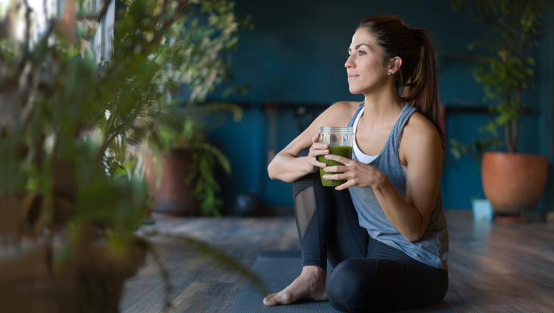 Female in workout attire sitting on yoga mat with green smoothie looking out a window.