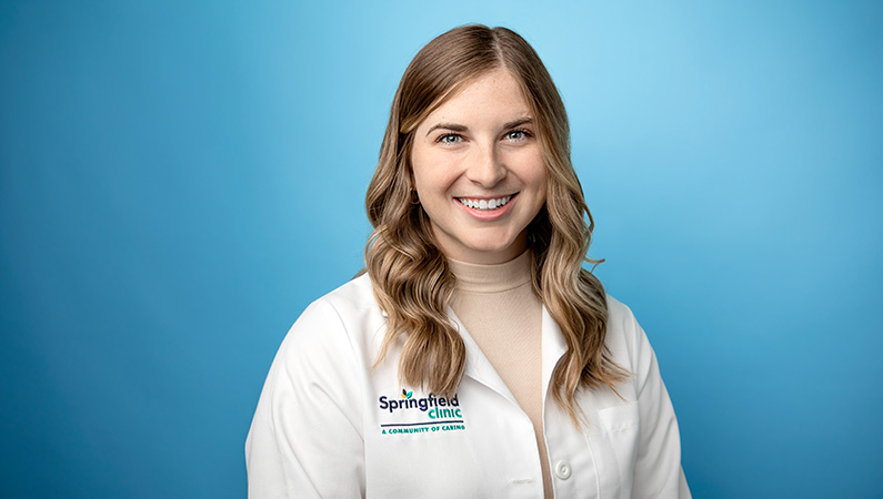 Female physician assistant in white lab coat smiling in front of a light blue photo backdrop.