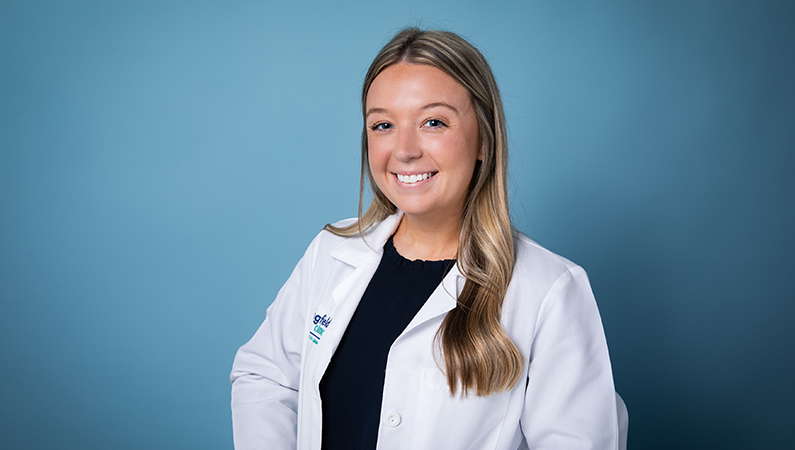 Female physician assistant in white coat smiling in front of a light blue backdrop.
