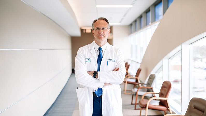 Male internal medicine doctor in white coat posing with arms crossed in a patient waiting room.
