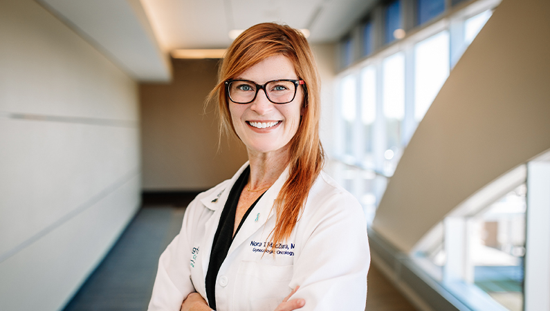 Female with red hair and glasses wearing white medical coat smiling in well-lit hallway