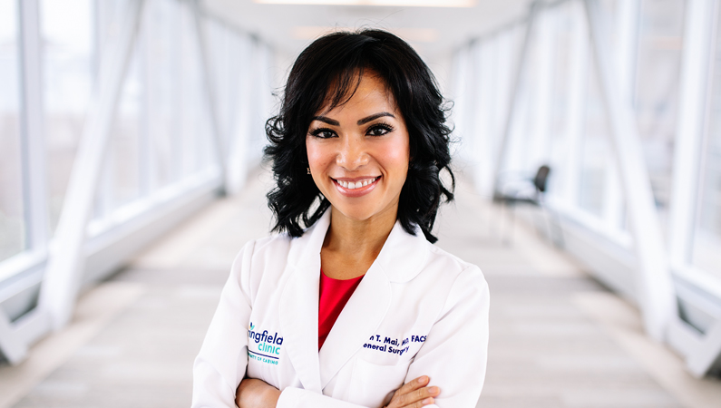 Female with short dark hair wearing white medical coat smiling in well-lit walkway