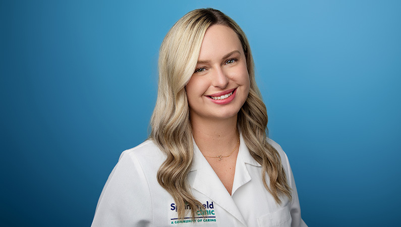 Female nurse practitioner wearing white medical coat smiling in front of light blue photo backdrop.