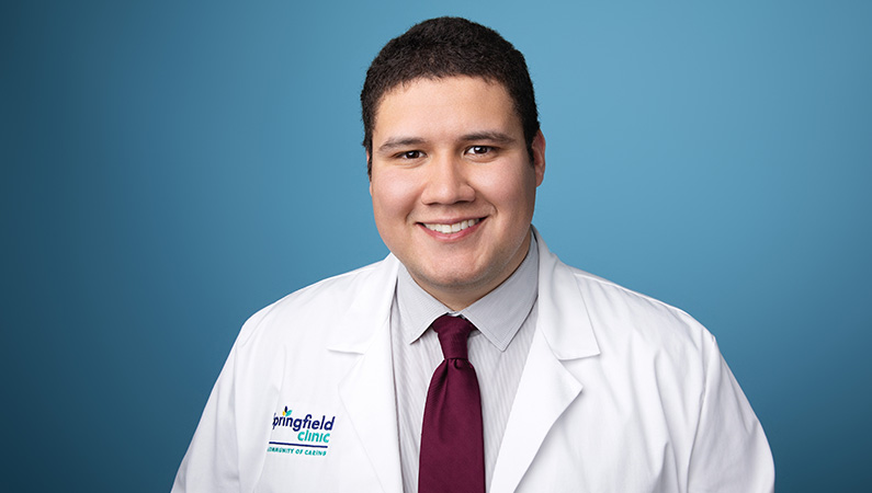 Male physician assistant wearing white medical coat smiling in front of blue photo backdrop.