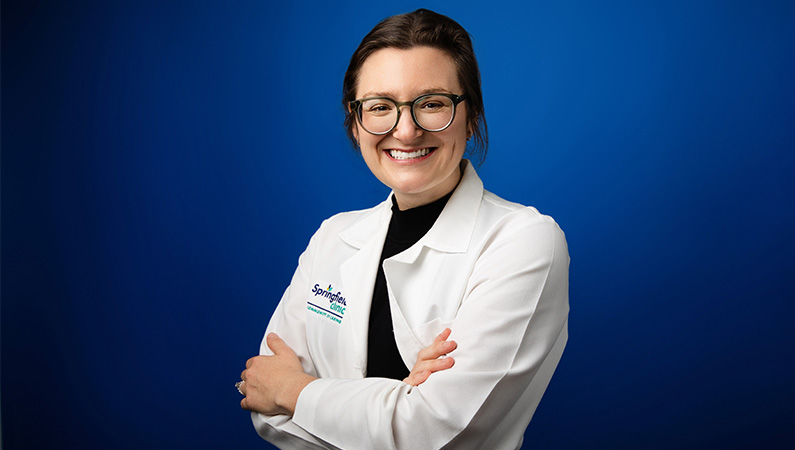 Female OB/GYN wearing white medical coat smiling in front of blue photo backdrop.