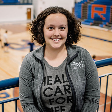 Female athletic trainer smiling with indoor basketball court in background.