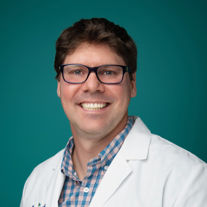 Male physician smiling in front of blue photo backdrop.