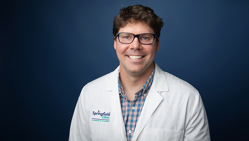 Male physician wearing white medical coat smiling in front of light blue photo backdrop.