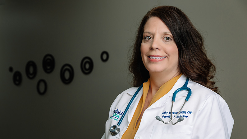 Female brunette nurse practitioner in white coat smiling in a dim office hallway setting.