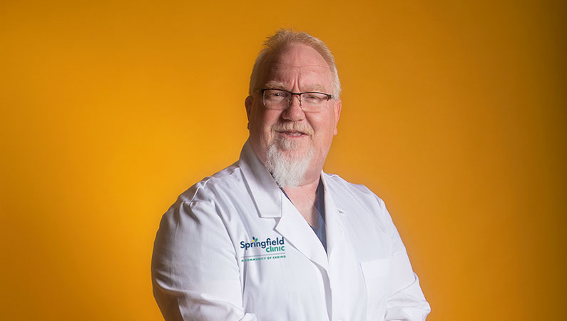 Male health care provider wearing white coat and glasses posing in front of a bright yellow backdrop