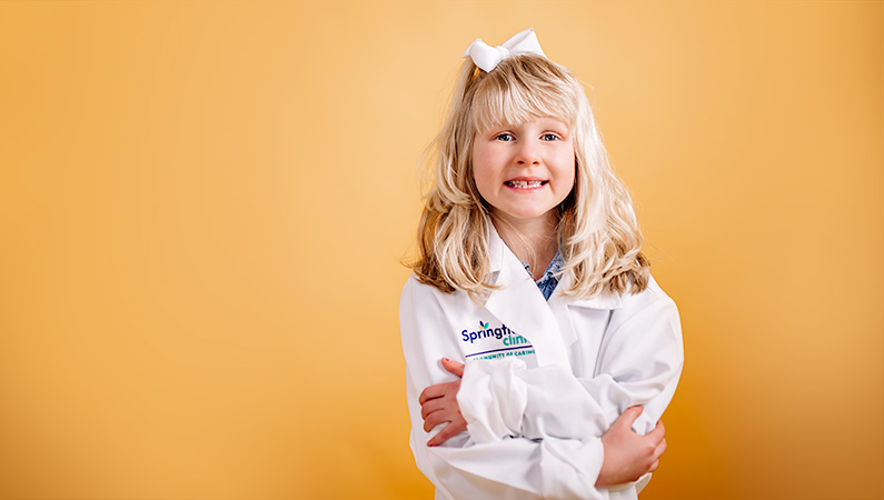 Child with blonde hair in oversized medical white coat crossing arms smiling on bright yellow background