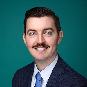 Male surgeon wearing suit smiling in front of green photo backdrop.