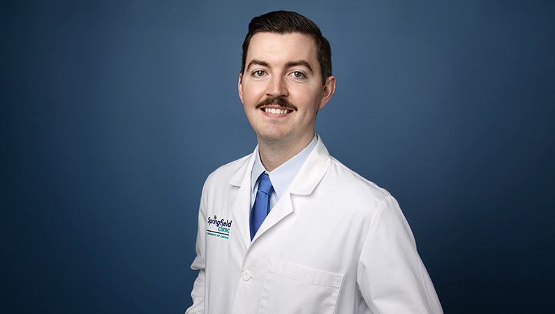 Male surgeon wearing white medical coat smiling in front of blue photo backdrop.