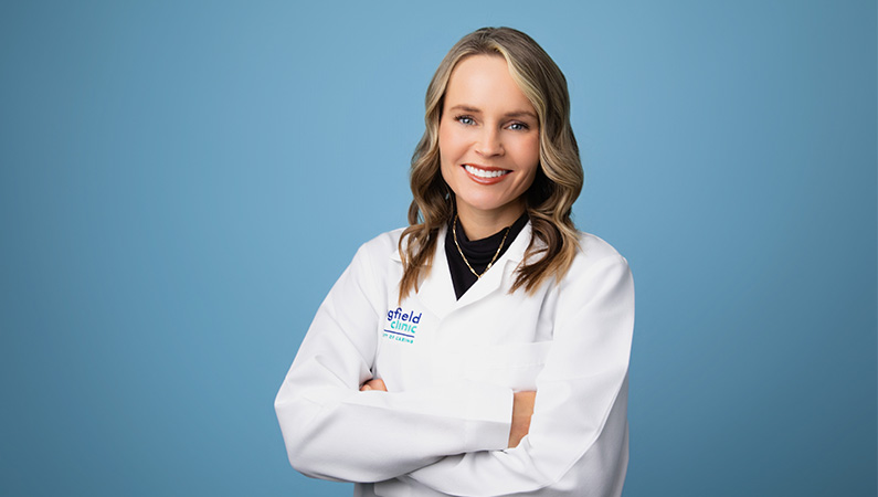 Female nurse practitioner in white medical coat smiling in front of blue photo backdrop.