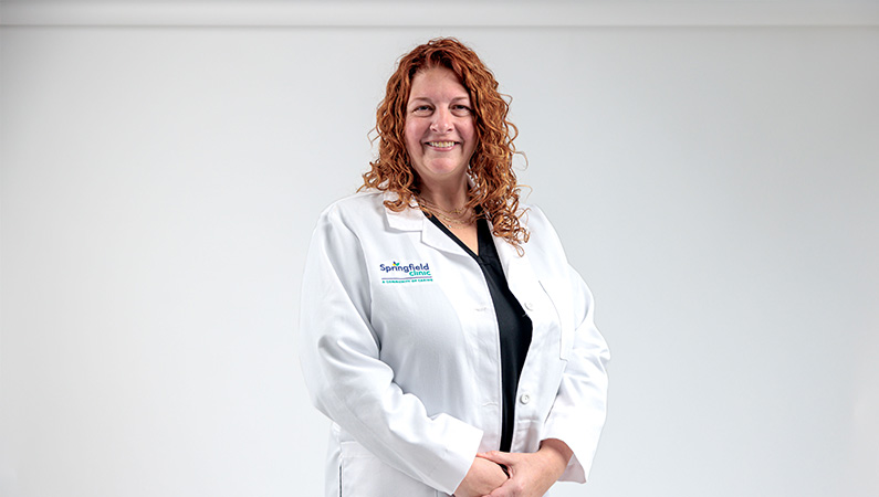 Female nurse practitioner in white medical coat smiling in front of a white photo backdrop.