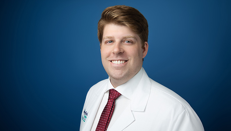 Male trauma surgeon wearing white medical coat smiling in front of light blue backdrop.