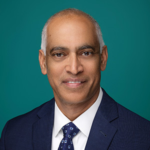 Male cardiologist smiling in professional headshot in front of teal blue backdrop.