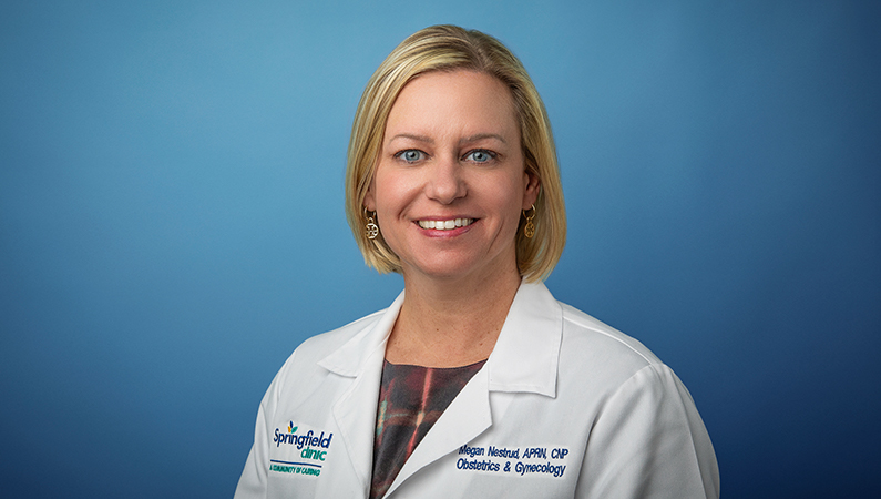 Female nurse practitioner wearing white medical coat smiling in front of light blue photo backdrop.
