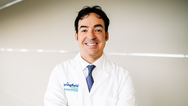 Male with dark hair wearing white medical coat smiling in well-lit setting