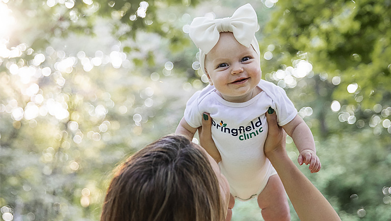 Baby being held up smiling wearing a white bow and onesie that says Springfield Clinic.