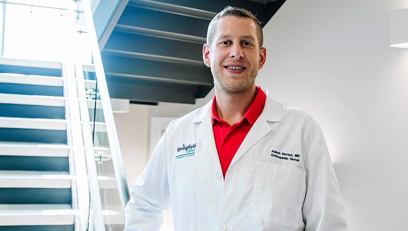 Man wearing a white doctors coat smiling in front of stairs.