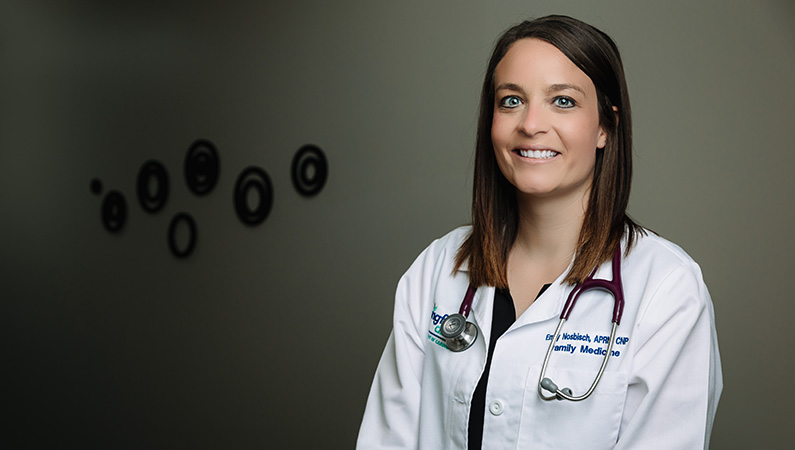 Female nurse practitioner in white coat smiling in a hallway.