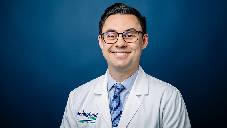 Male general surgeon wearing white medical coat smiling in front of light blue backdrop.