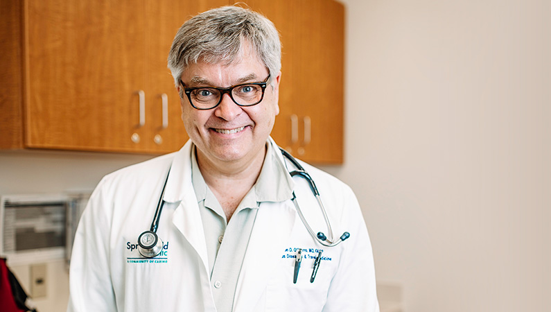 Man wearing a white doctors coat smiling in a well lit patient room.
