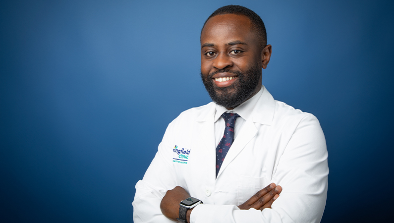 Male physician in white medical coat smiling in front of blue photo backdrop.