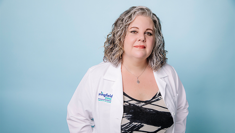 Female nurse practitioner wearing white coat posing in front of a light blue backdrop.