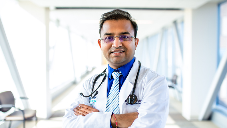 Male wearing glasses and white medical coat with stethoscope smiling in well-lit walkway