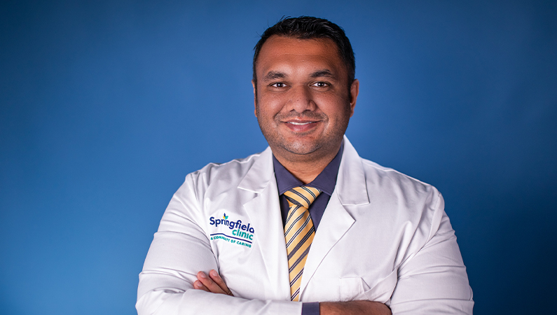 Male wearing white medical coat smiling with arms crossed on blue background