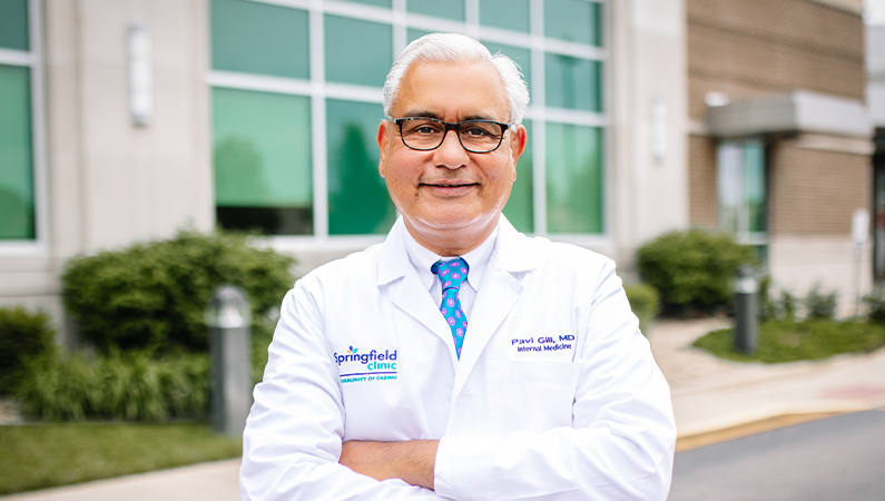 Male wearing glasses and white medical coat smiling in outdoor setting in front of medical building