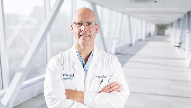 Male doctor in white coat crossing his arms in a glass hallway.