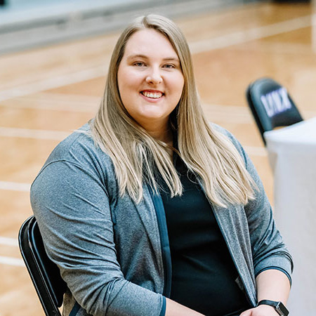 Female athletic trainer sitting in chair smiling in school gym setting.
