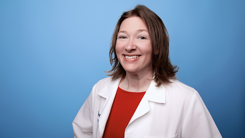 Female nurse practitioner in white medical coat smiling in front of blue photo backdrop.