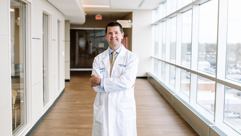 Male internal medicine doctor posing with arms crossed in a hallway with glass exterior windows.