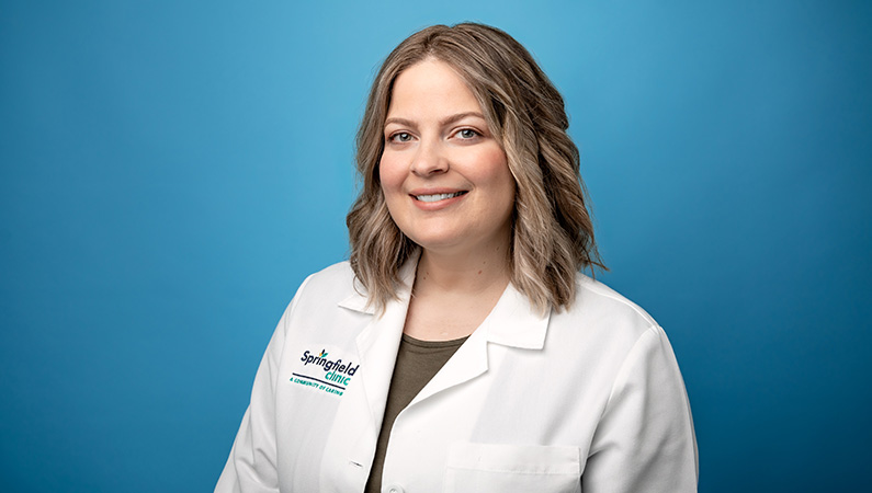 Female nurse practitioner in white medical coat smiling in front of light blue backdrop.
