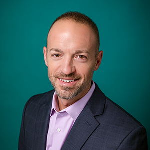 Male physician assistant in suit smiling in front of a teal blue backdrop.