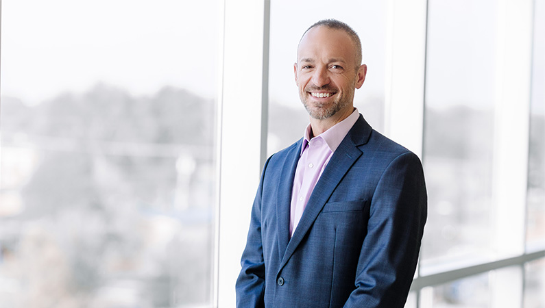 Male physician assistant in blue suit smiling in front of interior glass windows.