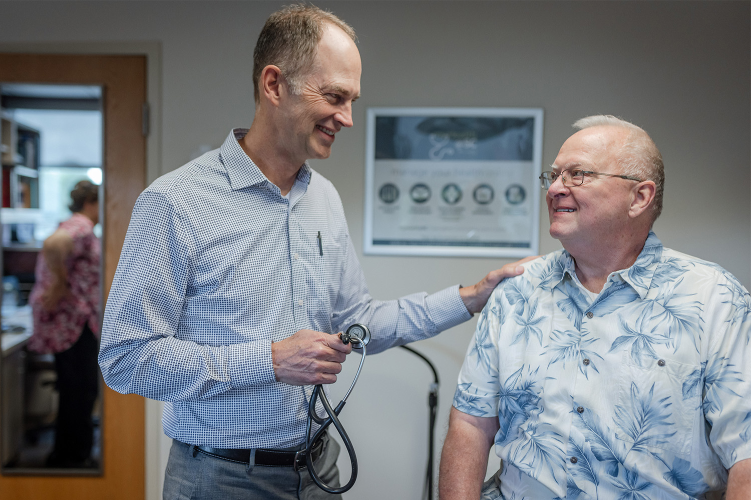 Doctor with stethoscope smiling while talking to elderly patient in an exam room.