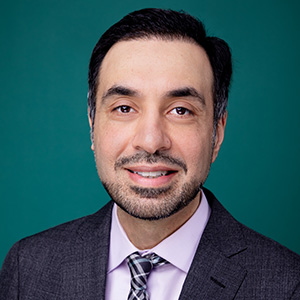 Male smiling in suit and tie in front of blue photo backdrop.